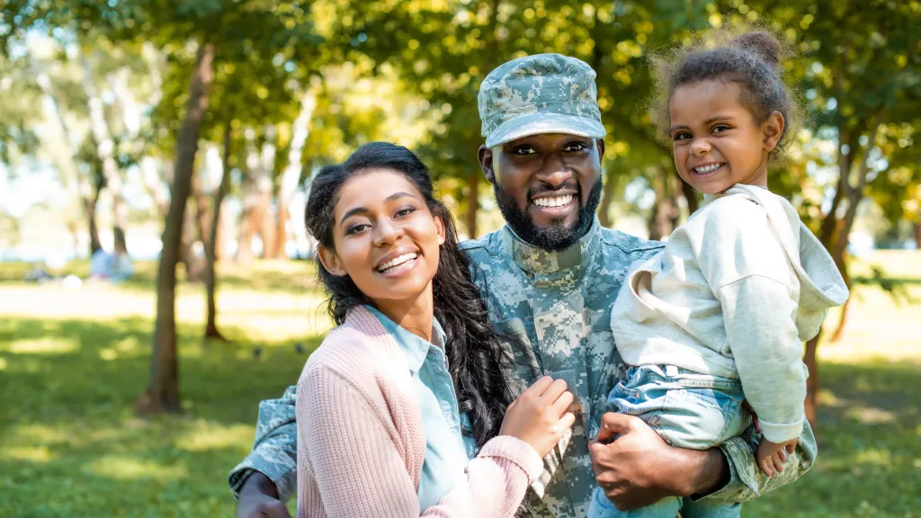 A man in a military uniform is standing outside with his family. He has one arm around his wife, and is holding his toddler-aged daughter in his other arm. Each family member is smiling.