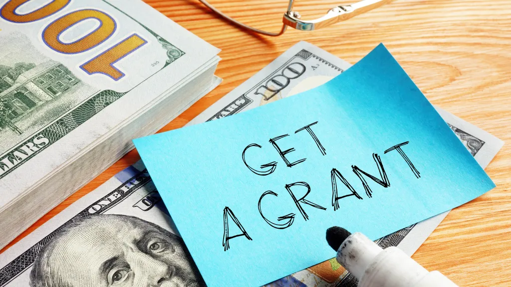 A note that says "Get a Grant" on top of a stack of 100 dollar bills.